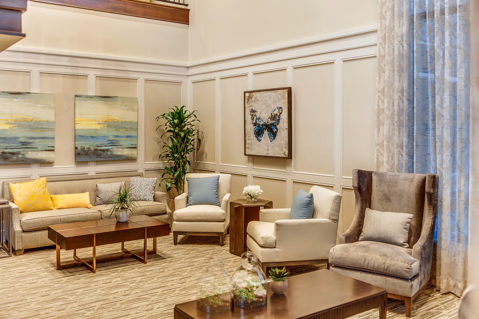 A cozy living room with a beige sofa adorned with yellow and gray pillows, two white armchairs with blue pillows, and a gray wingback chair with a matching pillow. The room features a wooden coffee table, side tables with plants and flowers, light-colored paneled walls, two abstract paintings, a butterfly artwork, and large patterned curtains.