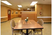 A small dining area with a wooden table and four beige cushioned chairs. The table has a small flower arrangement and a decorative runner. The room has beige walls and carpet, fluorescent ceiling lights, a fire extinguisher mounted on the wall, and a wooden door with a window in the background.