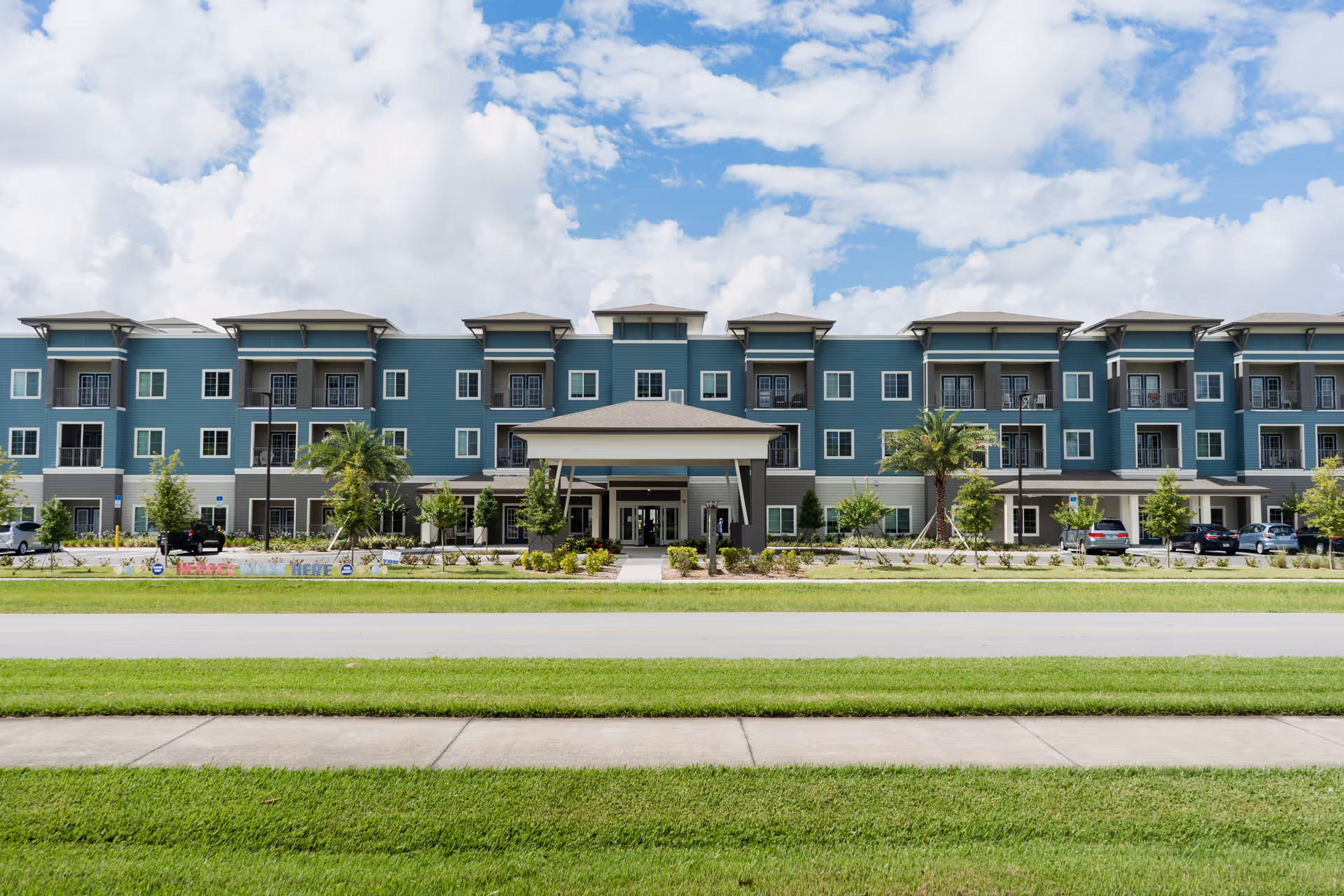 Front exterior of a three-story teal senior living building with a covered main entrance, balconies, and landscaped lawn.