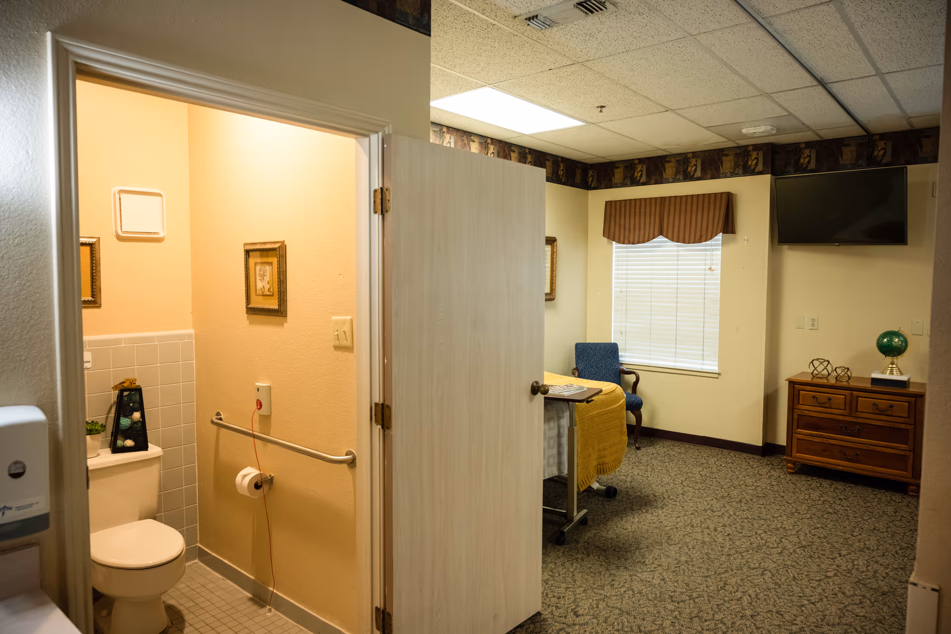 View of a room in a senior living facility showing a partially open door leading to a bathroom with a toilet, grab bar, and framed pictures on the wall. The main room has a window with blinds and a valance, a blue upholstered chair, a wooden dresser with decorative items, and a wall-mounted TV.