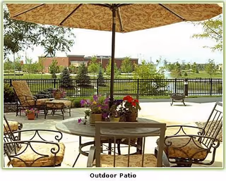 Outdoor patio area with a round table and cushioned chairs under a large umbrella. There are potted flowers on the table and lounge chairs nearby. In the background, there is a black metal fence, green trees, and a building visible across a grassy area.