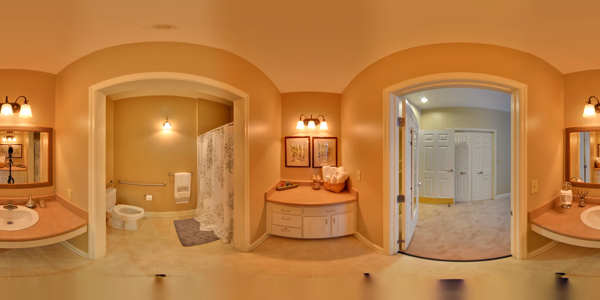 Panoramic view of a senior living facility bathroom with beige walls and tiled floor. The bathroom features two sinks with mirrors and light fixtures above them on either side. In the center, there is a toilet and a shower area with a curtain. To the right, an open doorway leads to a carpeted hallway with white doors.