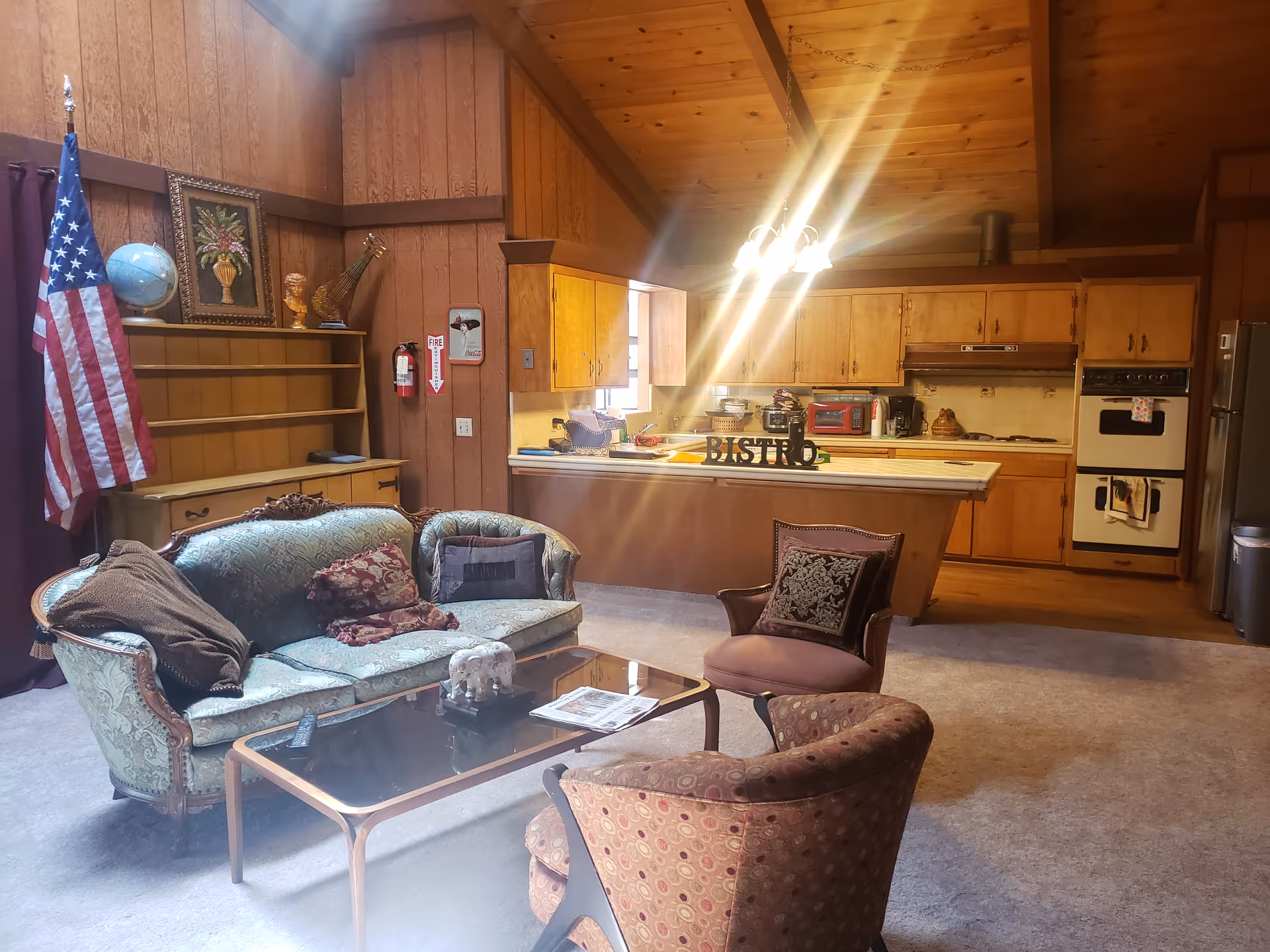 A cozy living area with vintage-style furniture including a green patterned sofa, two armchairs, and a glass coffee table. Behind the seating area is a wooden kitchen with cabinets, a stove, oven, and a countertop with a 'BISTRO' sign. The room has wood-paneled walls and a high wooden ceiling. An American flag and a globe are visible on a shelf to the left.