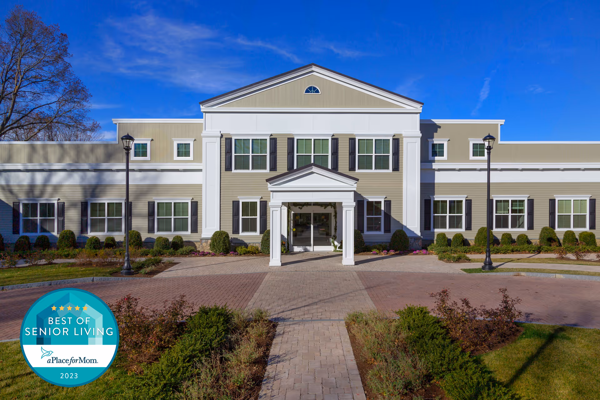 Front facade of a two-story senior living building with a paved driveway and landscaping, and a circular "Best of Senior Living" badge in the lower-left.