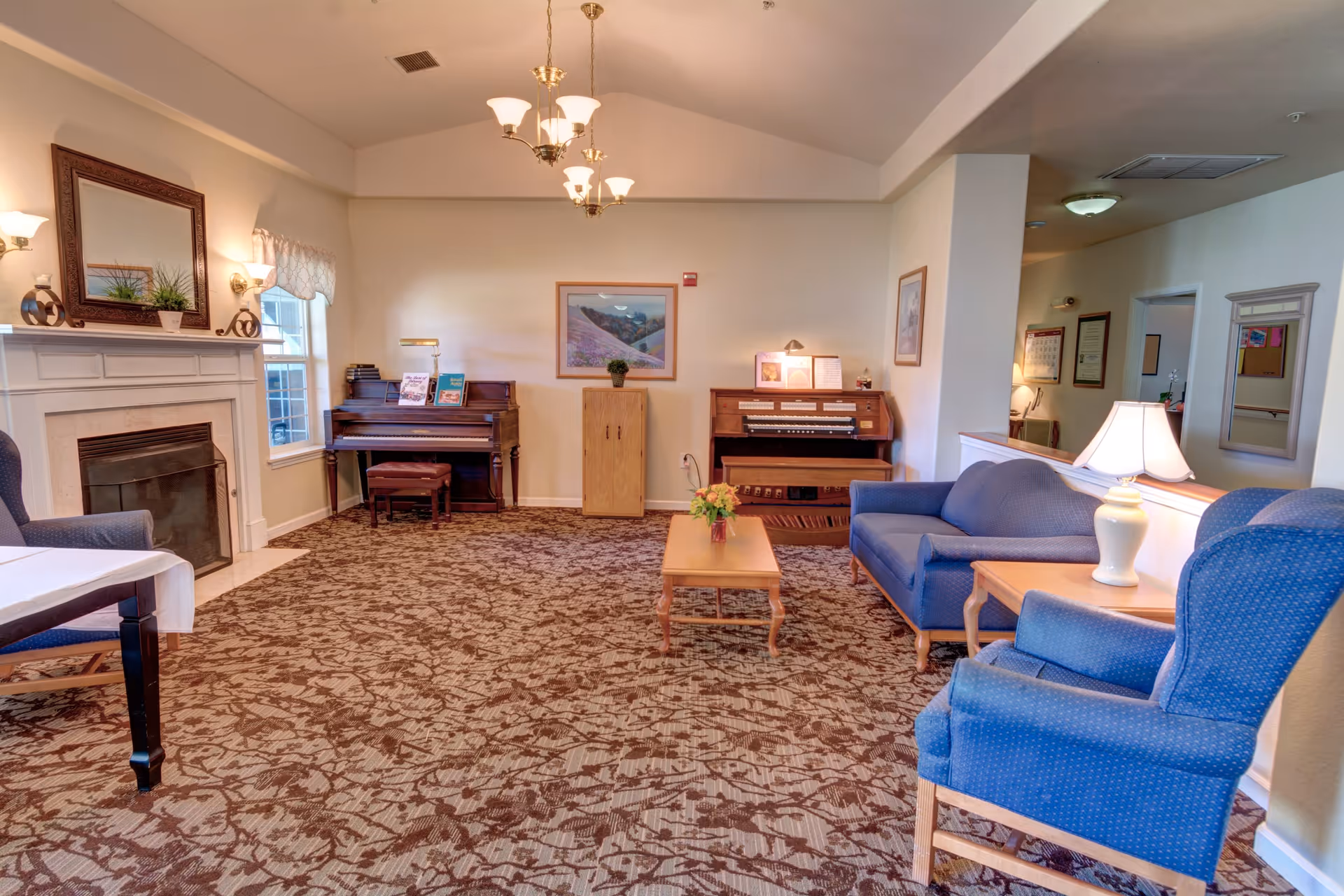 A cozy living room area in a senior living facility featuring a fireplace with a mirror above it, a piano and an organ against the far wall, a blue sofa and armchairs, a wooden coffee table with a flower arrangement, and a side table with a lamp. The room has patterned carpet and soft lighting from wall sconces and ceiling fixtures.
