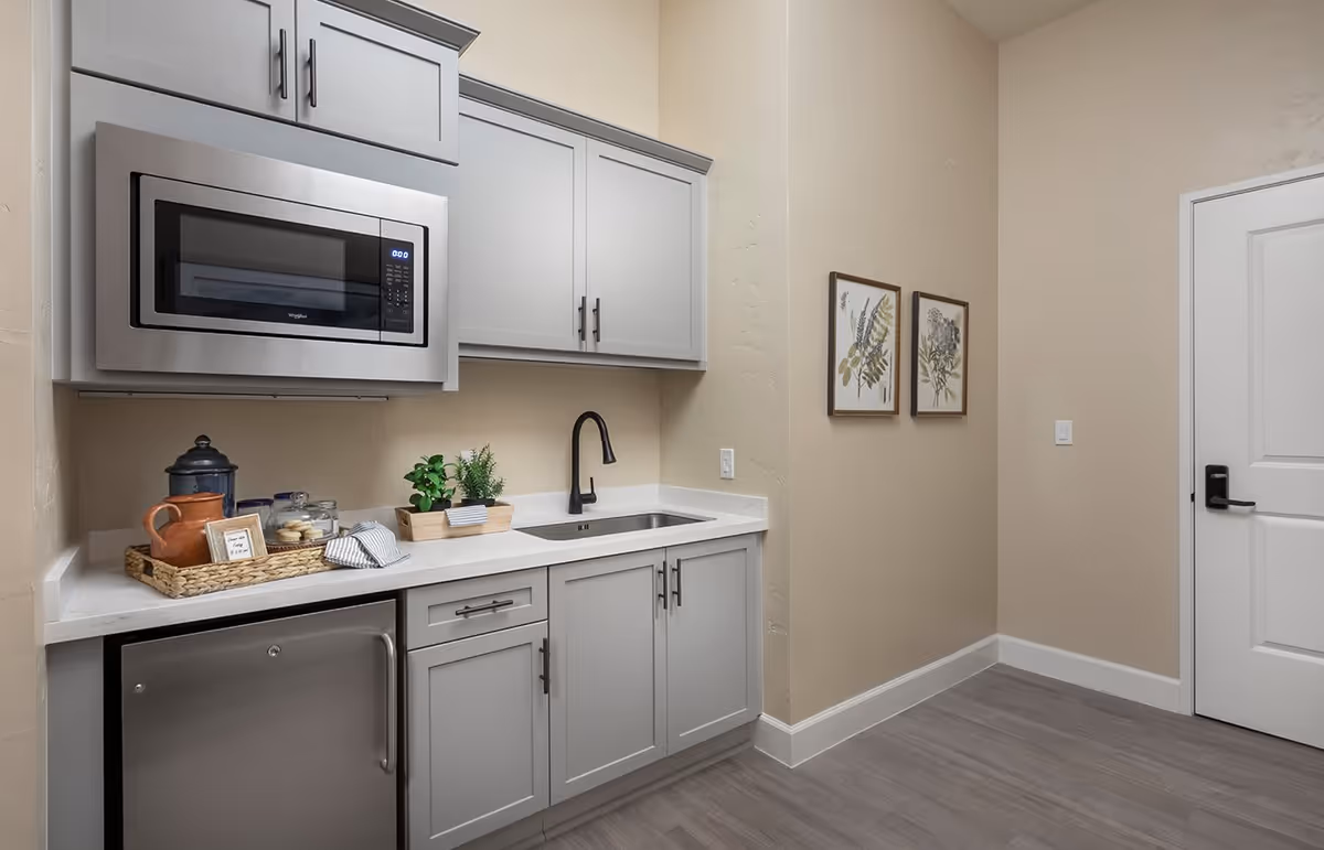 A small modern kitchen area with light gray cabinets, a stainless steel microwave mounted above the counter, a small stainless steel refrigerator below the counter, a black faucet over a stainless steel sink, and decorative items including a tray with jars and a small plant. The walls are beige with two framed botanical prints, and there is a white door with a black handle on the right side.
