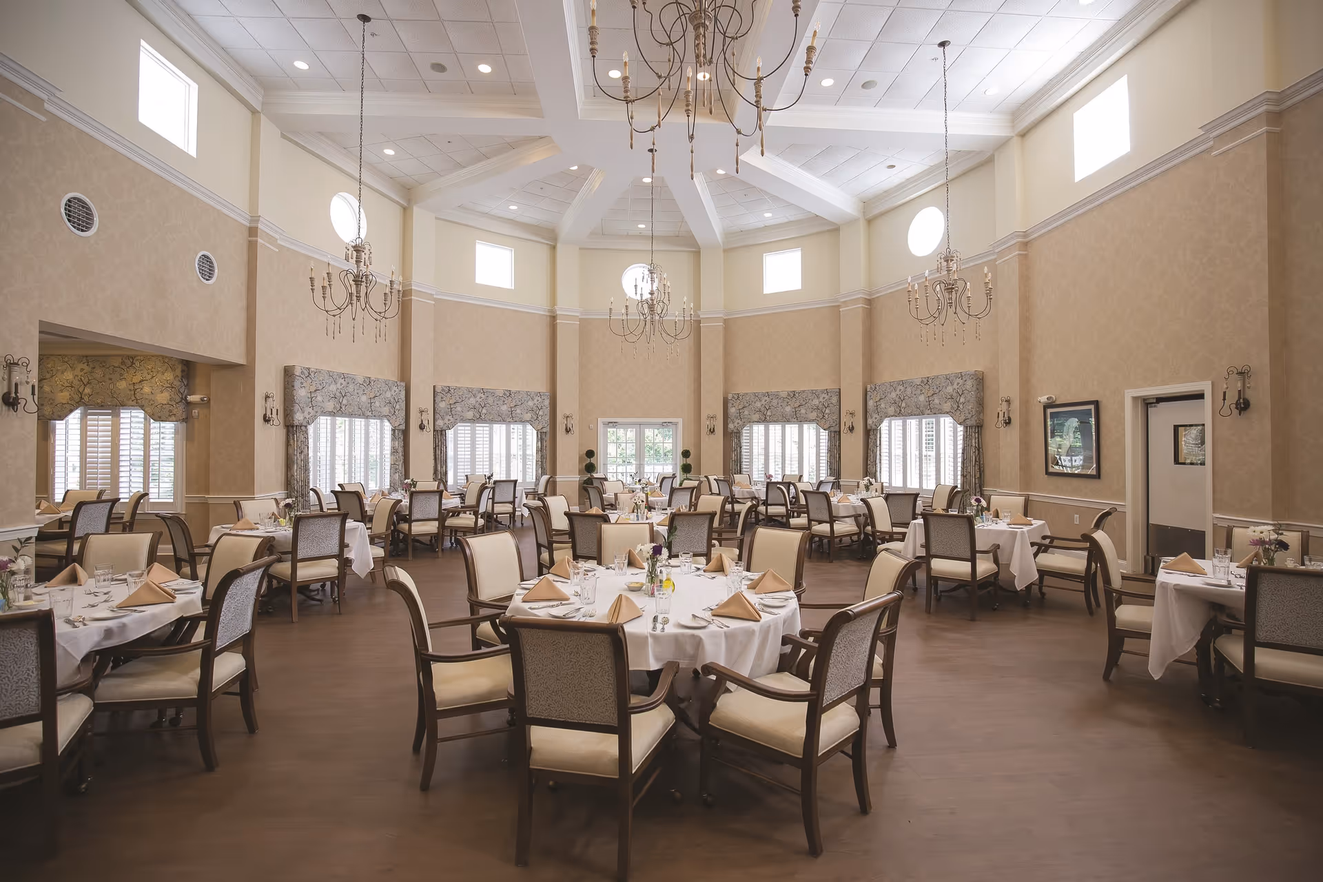 Spacious elegant dining room with round tables set with white linens, upholstered chairs, chandeliers, and high ceilings.