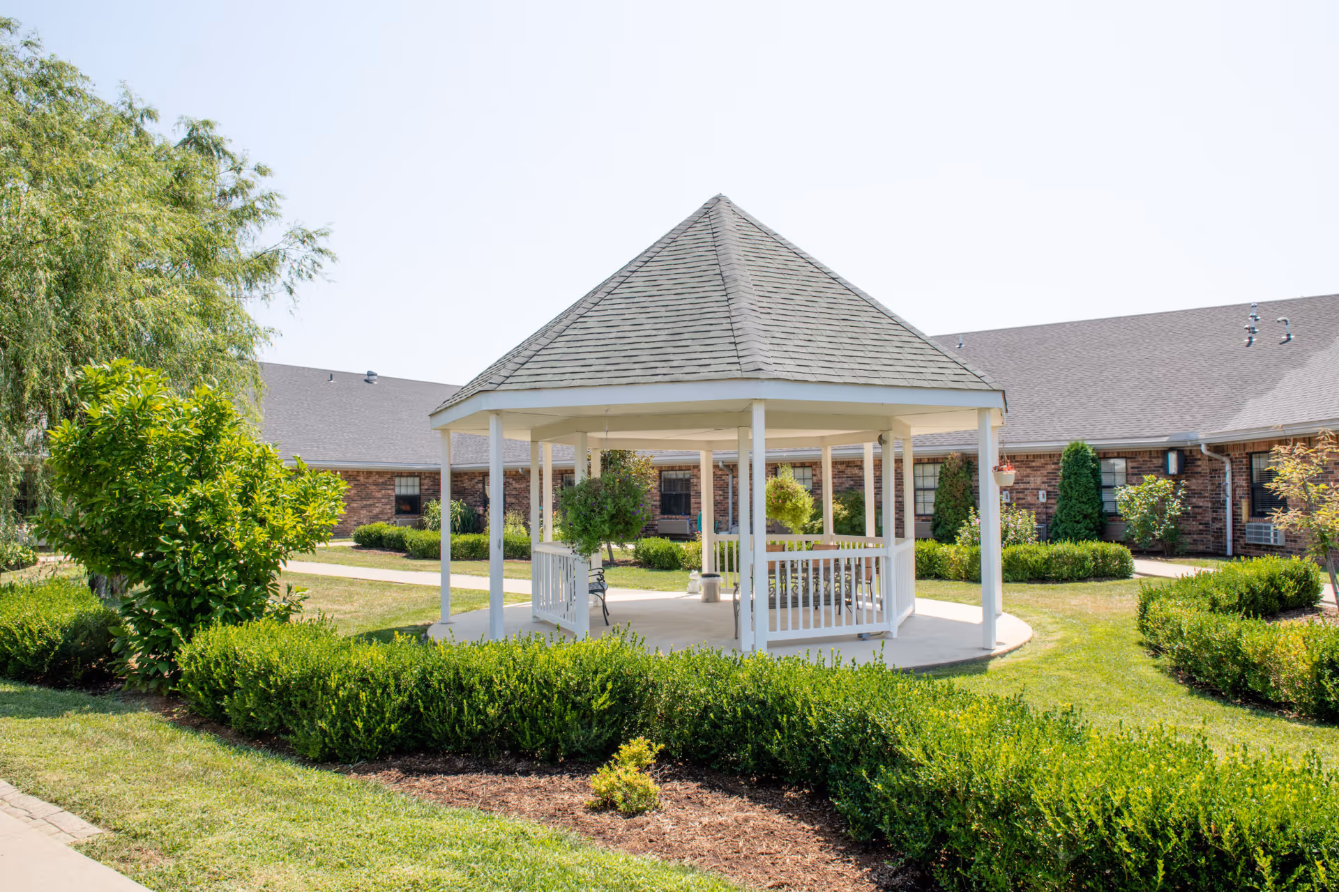 White octagonal gazebo in a landscaped courtyard surrounded by a brick senior living building and greenery.