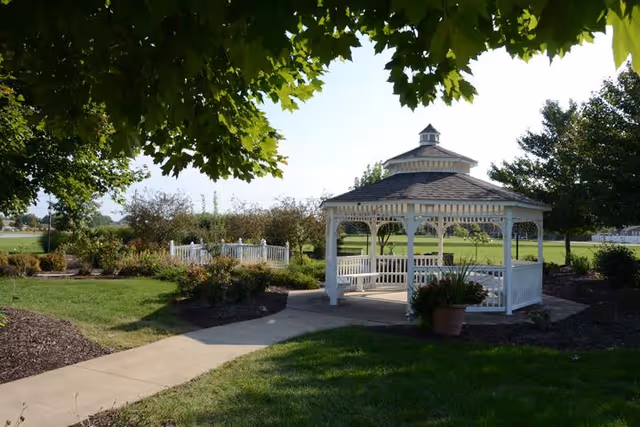 A white wooden gazebo with benches inside situated in a landscaped garden area with green grass, trees, and shrubs under a clear sky.