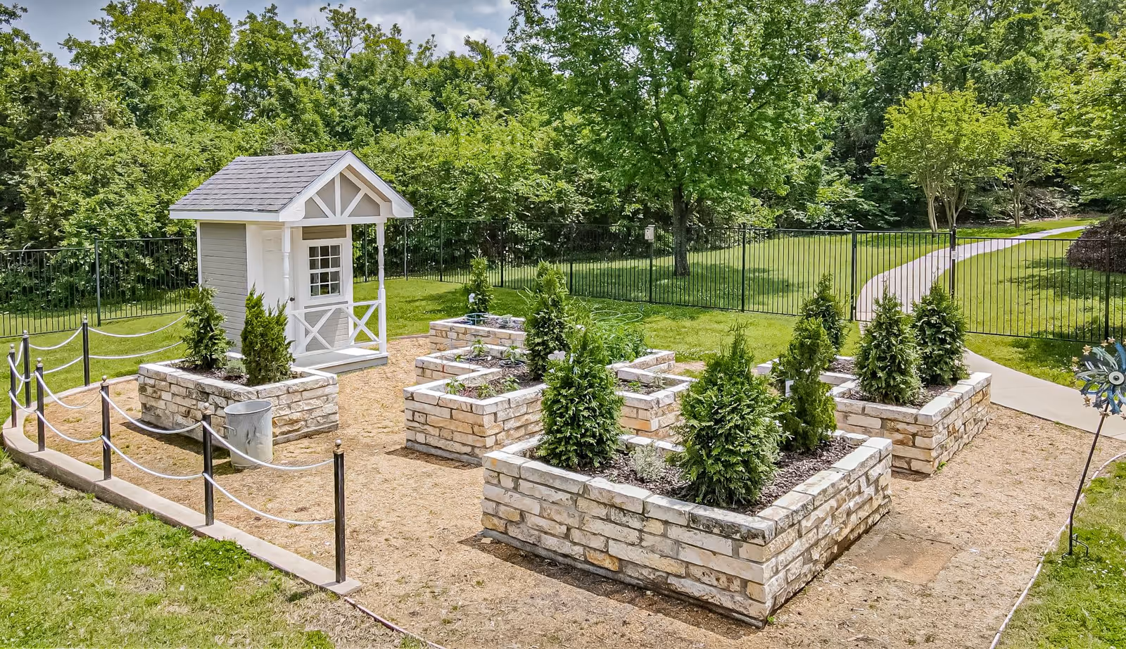 A small outdoor garden area with multiple raised stone planter beds containing small green shrubs and plants. There is a small white playhouse or garden shed with a gray roof on the left side. The garden is surrounded by a black metal fence and a paved pathway curves through the grassy area in the background under a partly cloudy sky.