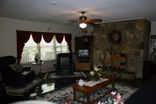 A cozy living room with a stone fireplace, a ceiling fan with light, two dark leather armchairs, a wooden coffee table with a floral centerpiece and a white teapot, floral rugs on the floor, and a window with burgundy curtains letting in natural light.