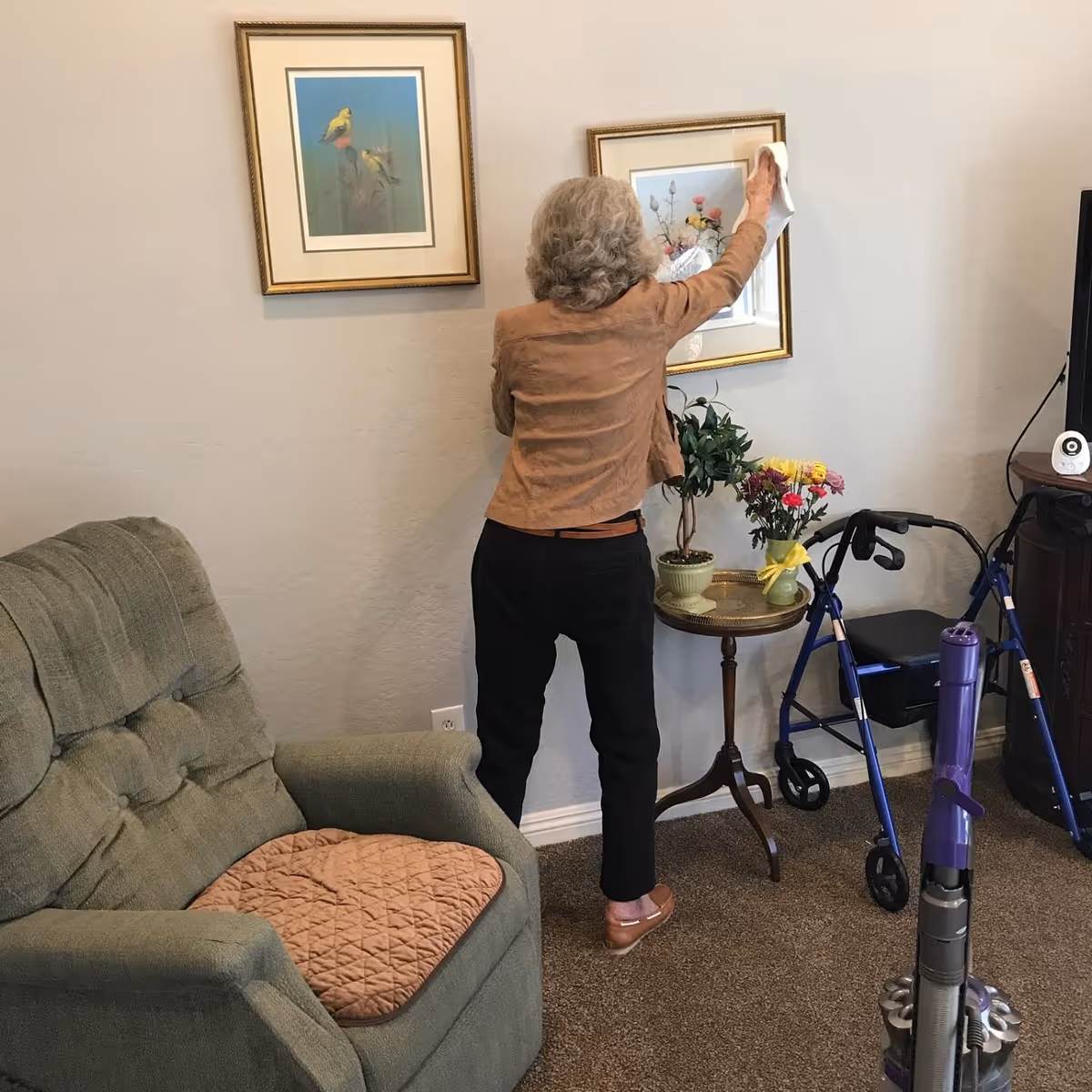 A person with gray hair wipes a framed picture on a wall in a living room, with a recliner, side table with flowers, a walker, and a vacuum in view.