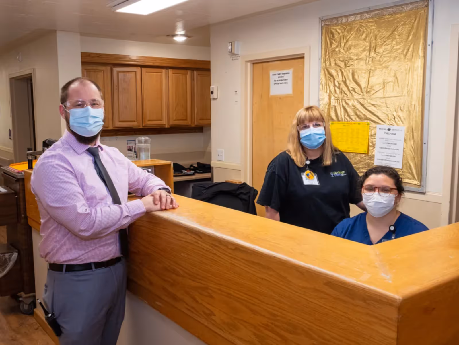 Three healthcare workers wearing face masks standing behind and in front of a wooden reception desk in a healthcare facility. The background shows wooden cabinets and a door with notices posted on it.
