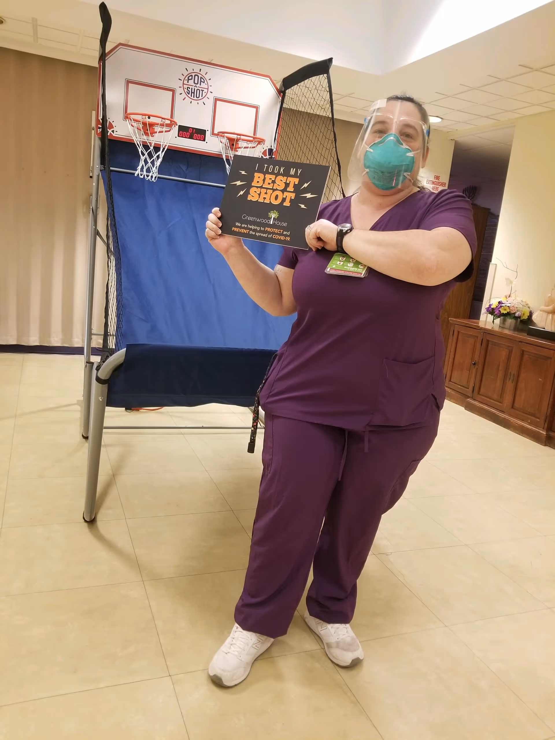A healthcare worker wearing purple scrubs, a green face mask, and a clear face shield stands indoors holding a sign that reads 'I TOOK MY BEST SHOT' with the Greenwood House logo and a message about protecting and preventing the spread of COVID-19. Behind her is a Pop-A-Shot basketball arcade game and some wooden furniture with flowers on top.