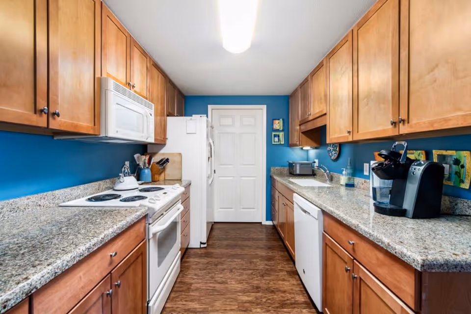A modern kitchen with wooden cabinets, granite countertops, and blue walls. The kitchen features a white electric stove with a microwave above it, a white refrigerator, a dishwasher, a coffee maker, a toaster, and various kitchen utensils. The floor is wooden, and there is a white door at the end of the kitchen.