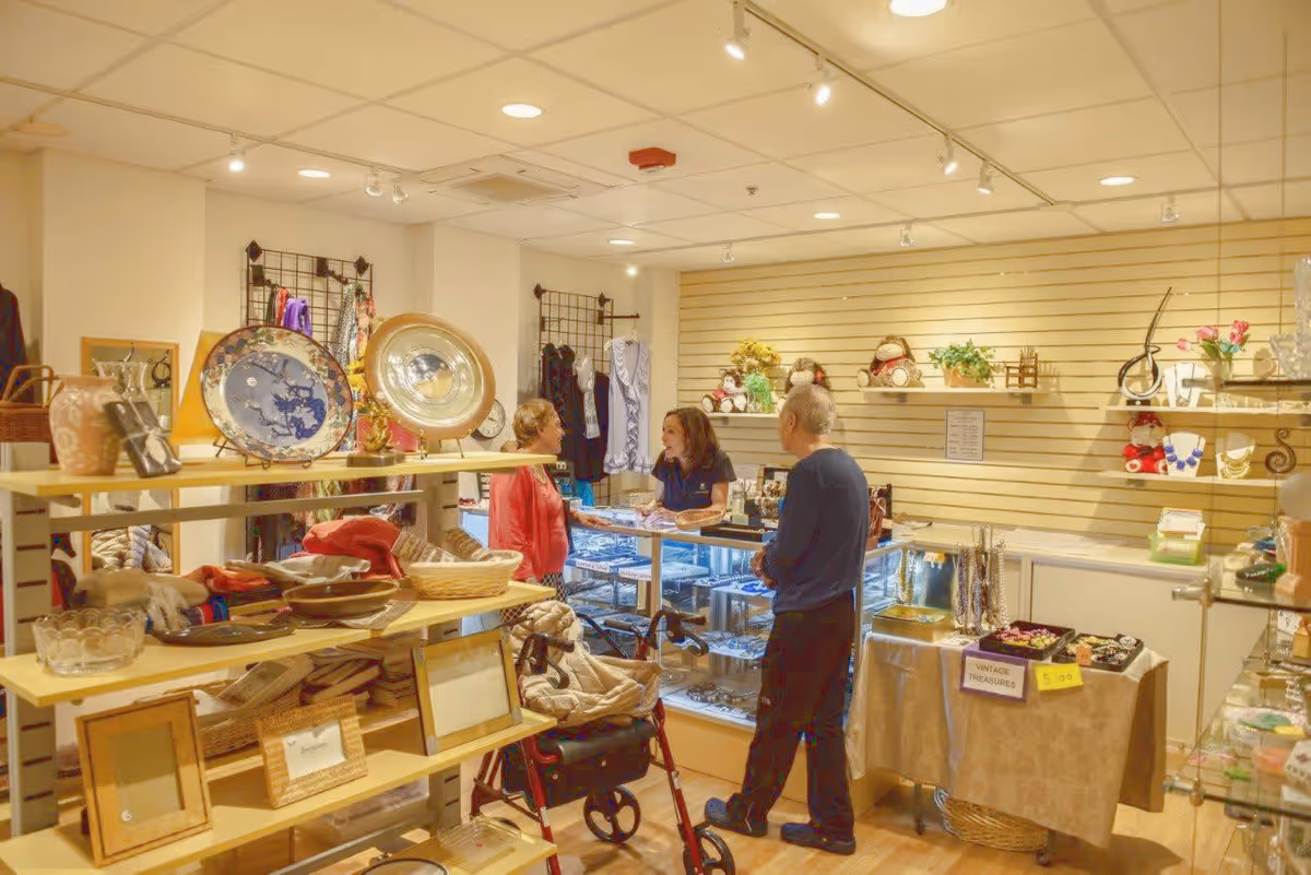 Interior of a small gift shop or boutique within a senior living facility. Shelves display various items including picture frames, decorative plates, scarves, and jewelry. Three people are present: a woman behind the counter talking to an elderly woman and an elderly man standing nearby with a walker. The walls have slat panels with small decorative items and plants.