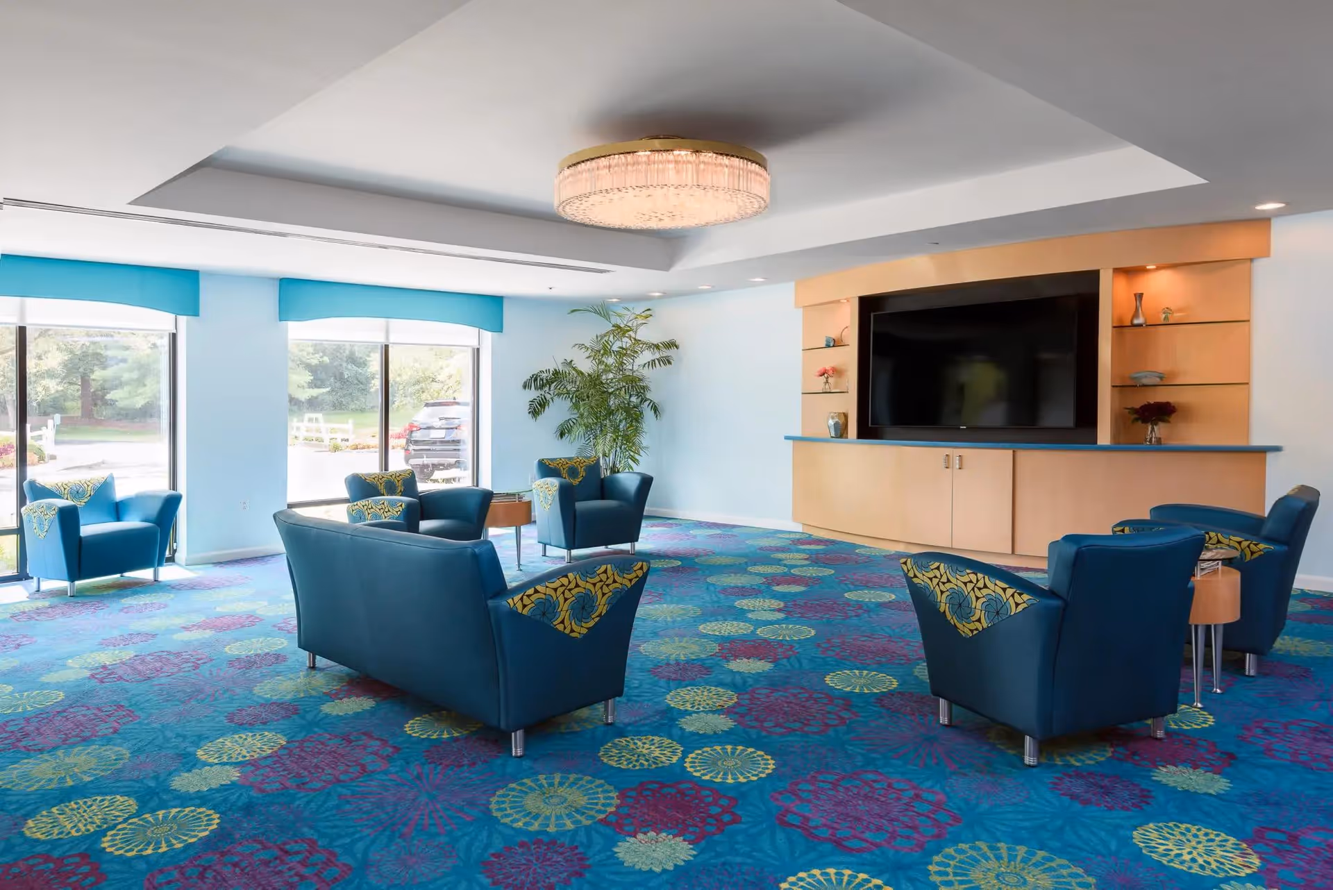 Bright common lounge with teal armchairs arranged around a large TV and a colorful patterned carpet.