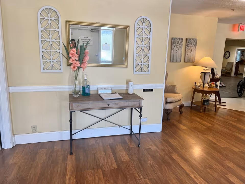 Interior view of a senior living facility hallway with wooden flooring. A small table against a beige wall holds a vase with pink flowers, a bottle of hand sanitizer, and a visitor sign-in book. Above the table is a rectangular mirror flanked by two decorative white wall hangings. To the right, there is a cozy seating area with a beige armchair, a wooden side table with a lamp, and wall art. In the background, a wheelchair and a clock are visible in another room.