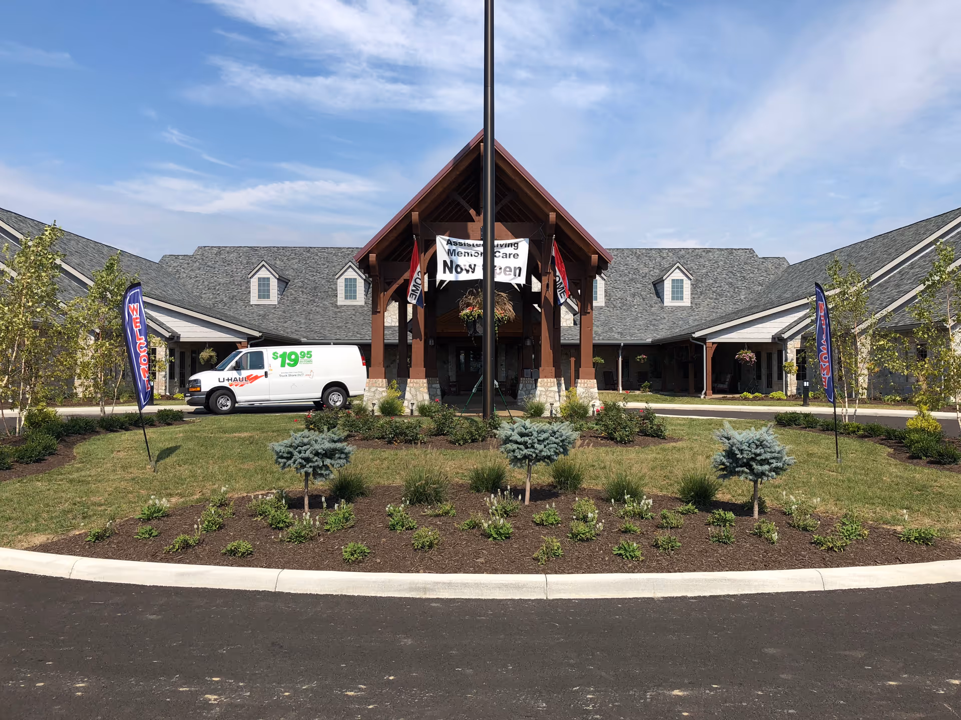 Front exterior view of the Inn at Apple Ridge facility with a landscaped circular driveway, a U-Haul van parked on the left side, and banners that say 'WELCOME'. The building has a peaked roof and a covered entrance with a sign indicating 'Memory Care Now Open'.