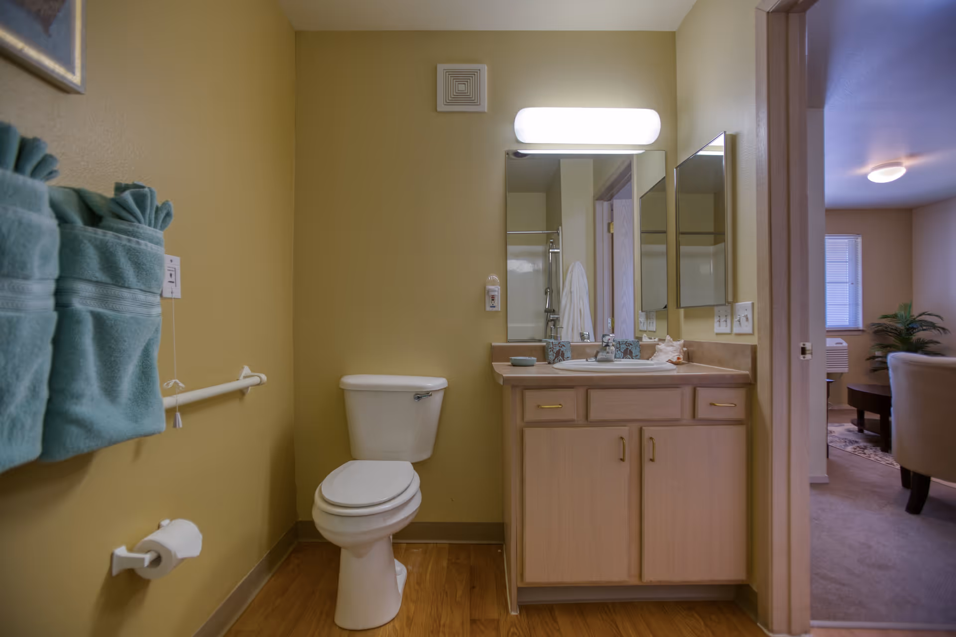 A bathroom with a toilet, a vanity with a sink, and a mirror above it. There are three teal towels hanging on a towel rack on the left wall. The bathroom has light brown walls and wood flooring. Through an open doorway, a living room area with a chair, a window, and a plant is visible.