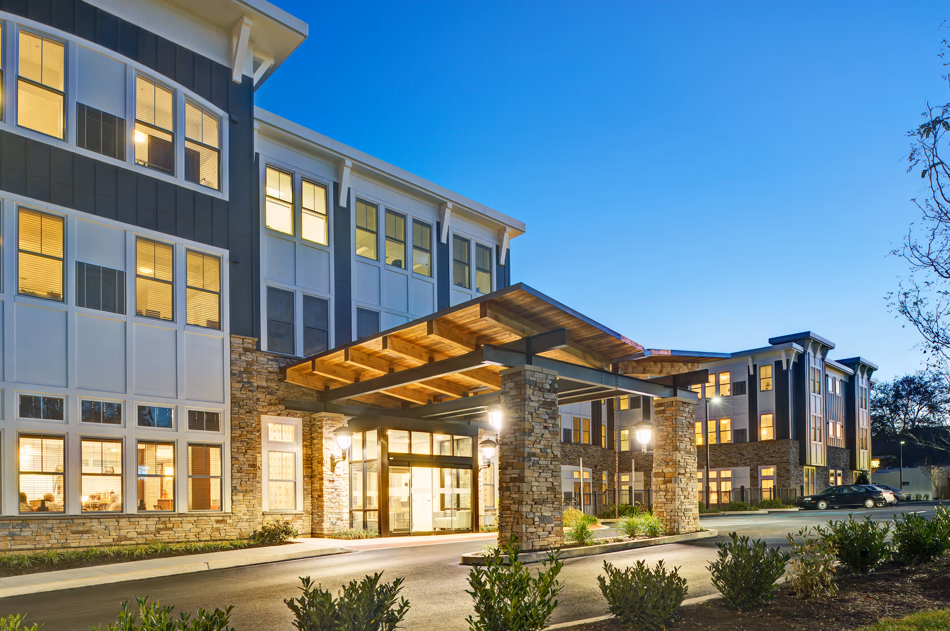 Exterior view of a modern senior living facility building at dusk with large windows illuminated from inside, stone pillars supporting a wooden canopy over the entrance, and a driveway with parked cars.