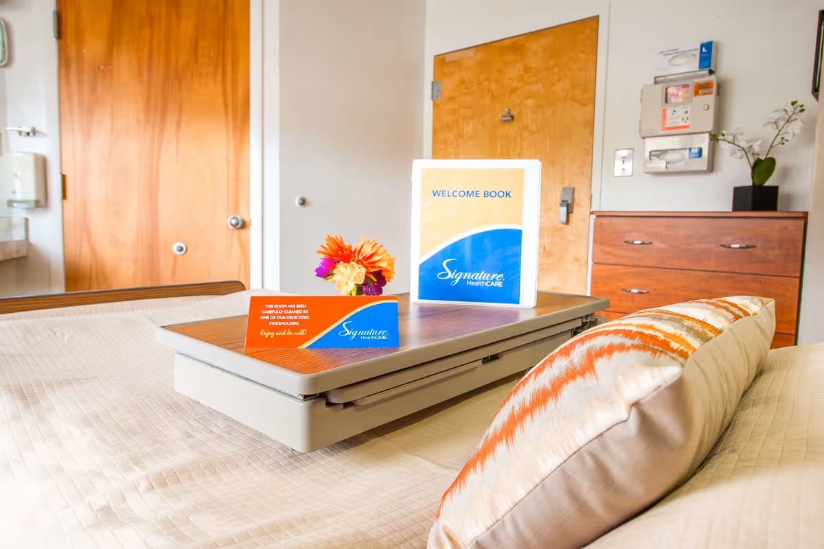 A neatly made bed with a beige quilt and a decorative pillow with orange and white patterns in a room. On the bed is a tray holding a small bouquet of flowers, a welcome book, and a card indicating the room has been carefully cleaned. In the background, there are wooden doors, a wooden dresser with a small plant, and a wall-mounted dispenser.