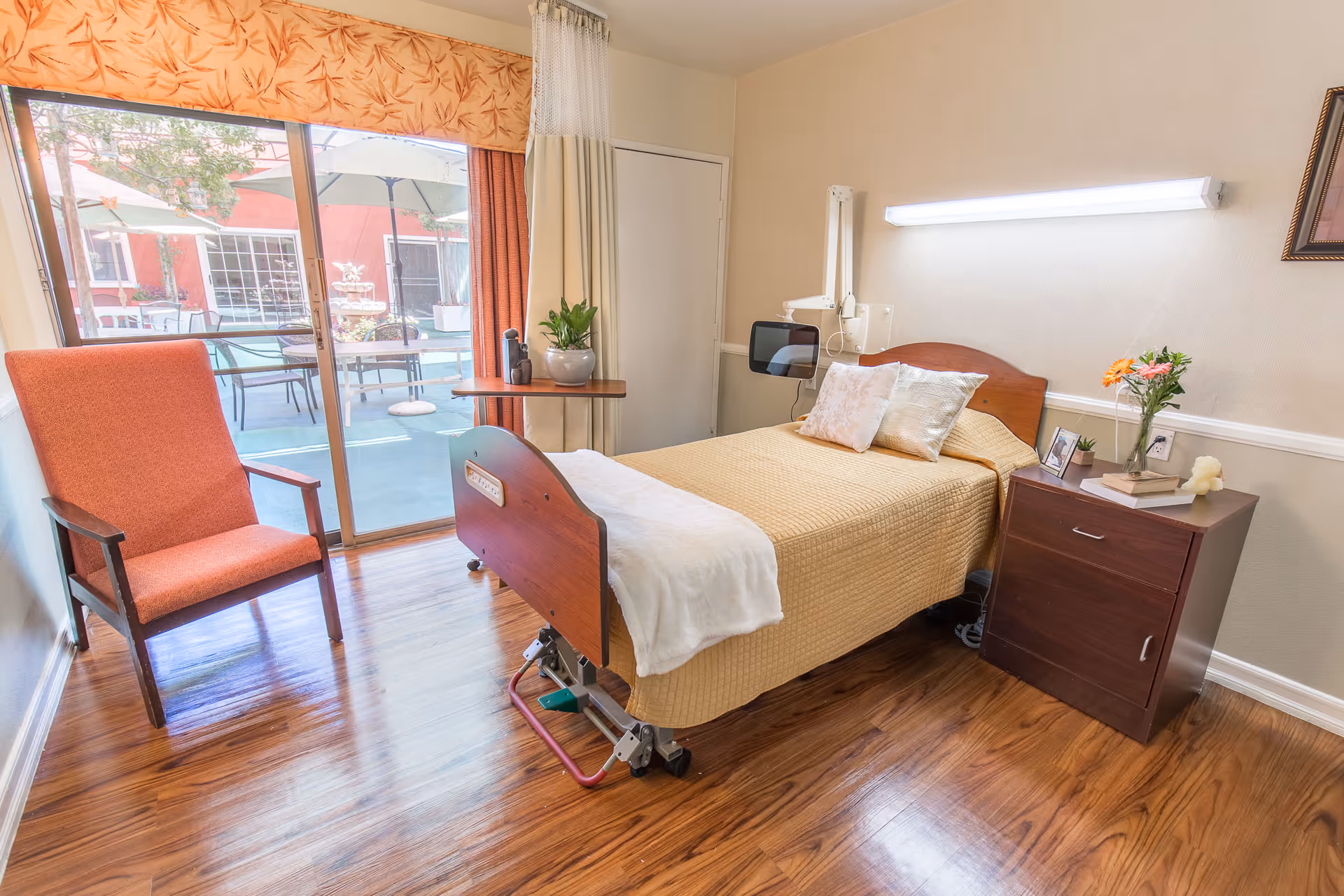 A bright and clean senior living bedroom with a single bed covered in a beige quilt and white blanket. The room has wooden flooring, a wooden nightstand with flowers, books, and a photo frame, and an orange armchair near a sliding glass door that opens to an outdoor patio with tables, chairs, and umbrellas.
