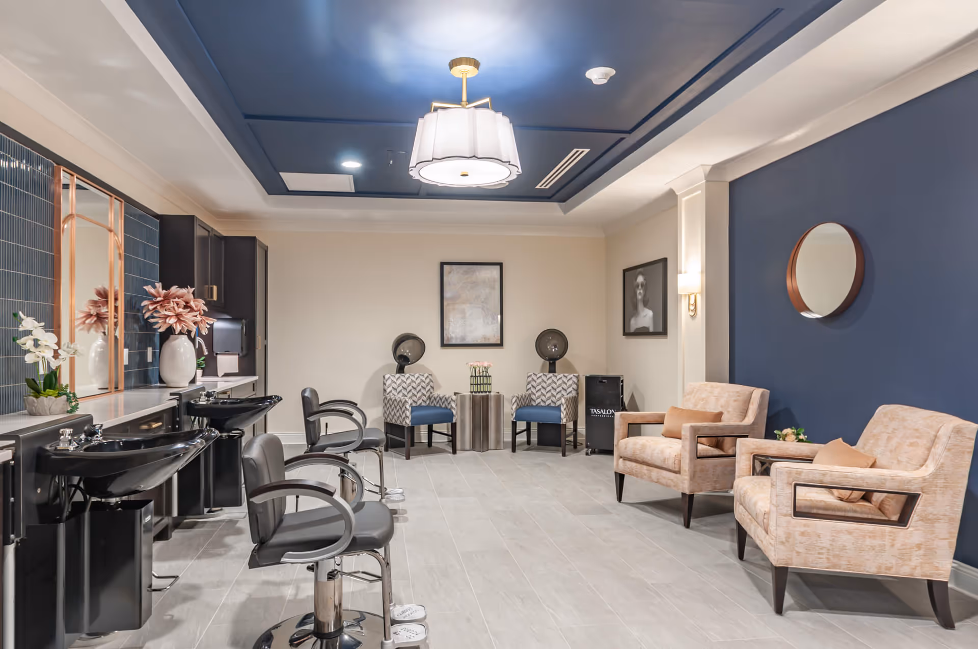 Interior view of a modern salon area with two black hair washing stations and two black salon chairs on the left. The back wall has two patterned armchairs with blue cushions and a small round table with flowers between them. On the right, there are two beige armchairs with brown pillows and a round mirror on a dark blue accent wall. The ceiling has a blue recessed section with a white hanging light fixture.