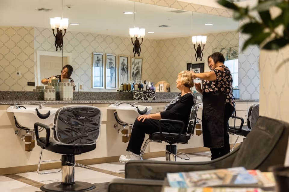 A hair salon area inside The Park at Surprise Independent Living Community, showing a hairstylist cutting an elderly woman's hair. The room features salon chairs, wash basins, a large mirror, patterned wallpaper, and soft lighting fixtures.