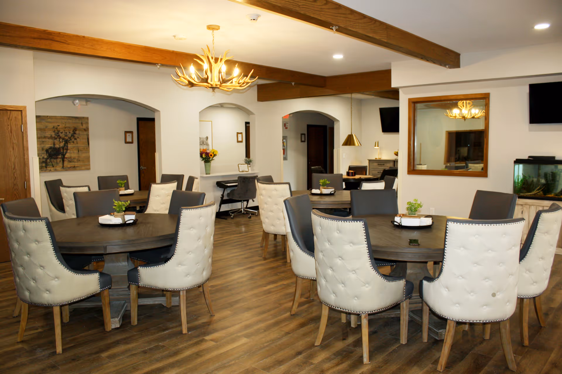 A well-lit dining room with multiple round wooden tables surrounded by cushioned chairs in white and gray. The room features wooden beams on the ceiling, a chandelier with an antler design, a large mirror on the wall, a fish tank, and decorative artwork including a deer painting. The floor is wood, and there are small plants and napkins on the tables.