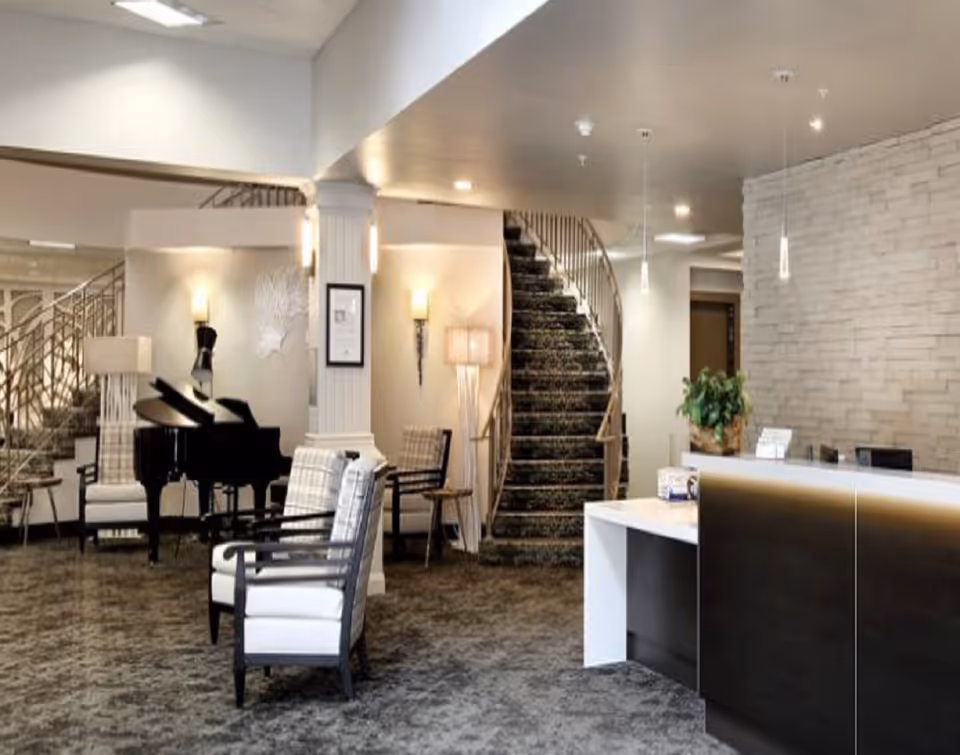 A modern and elegant senior living facility lobby featuring a curved staircase with carpeted steps and metal railing, a black grand piano, several cushioned armchairs with plaid upholstery, a reception desk with a dark front panel and white countertop, decorative wall sconces, and a stone accent wall behind the reception area.