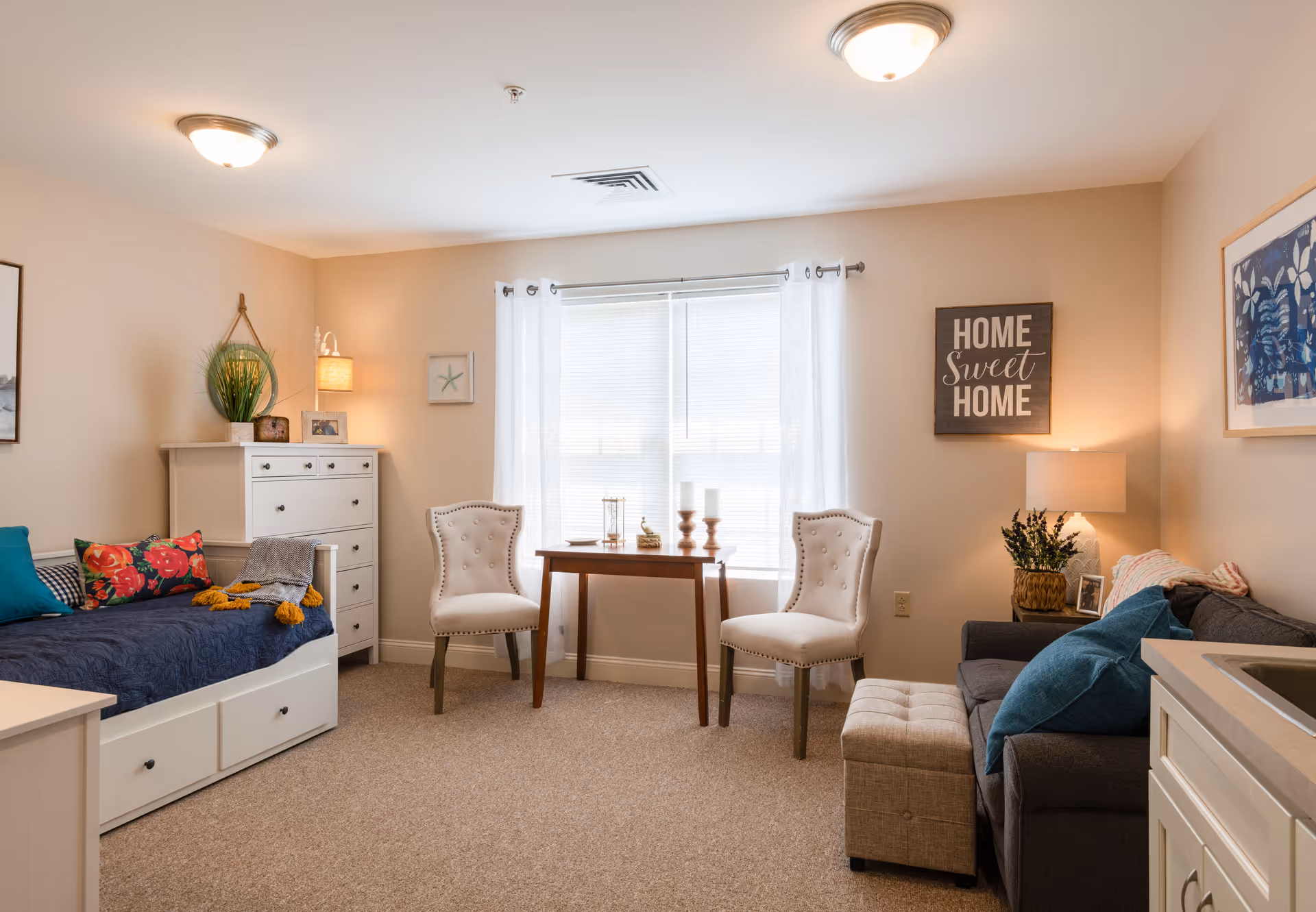 A cozy living room area with a white daybed adorned with colorful pillows and a gray throw blanket on the left. Next to it is a white chest of drawers with a potted plant and framed photos on top. In front of a window with white curtains, there is a small wooden table with two white upholstered chairs. On the right side, a gray sofa with blue pillows and a beige ottoman is placed next to a side table with a lamp and a plant. The walls are decorated with framed artwork and a sign that reads 'HOME Sweet HOME'.