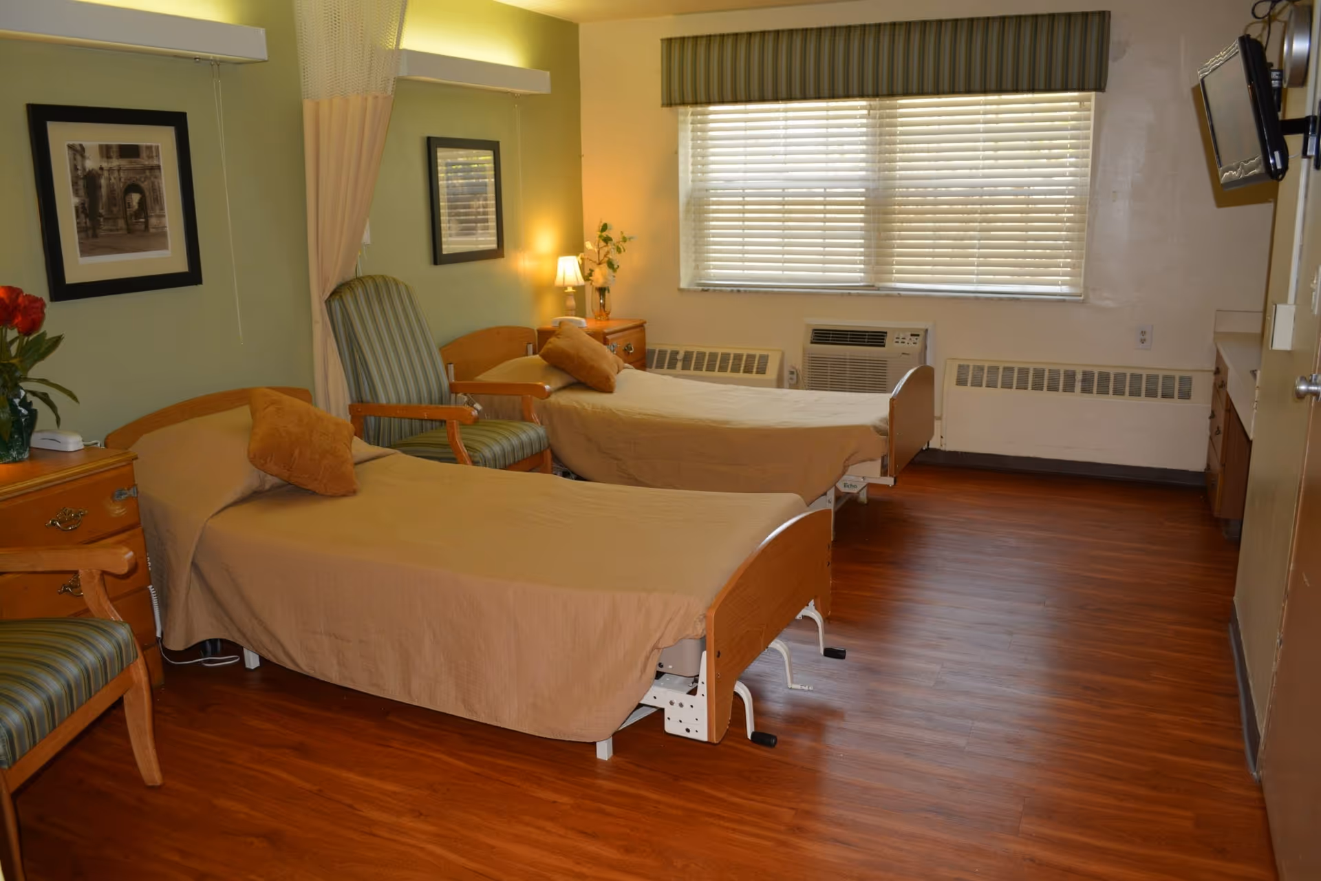 A nursing facility room with two single beds covered in beige bedspreads, each with a pillow. Between the beds is a small wooden nightstand with a lamp and a vase with flowers. There are two striped armchairs, one near the nightstand and one in the foreground. The room has wooden flooring, a large window with blinds and a valance, and framed pictures on the wall. An air conditioning unit is installed below the window.