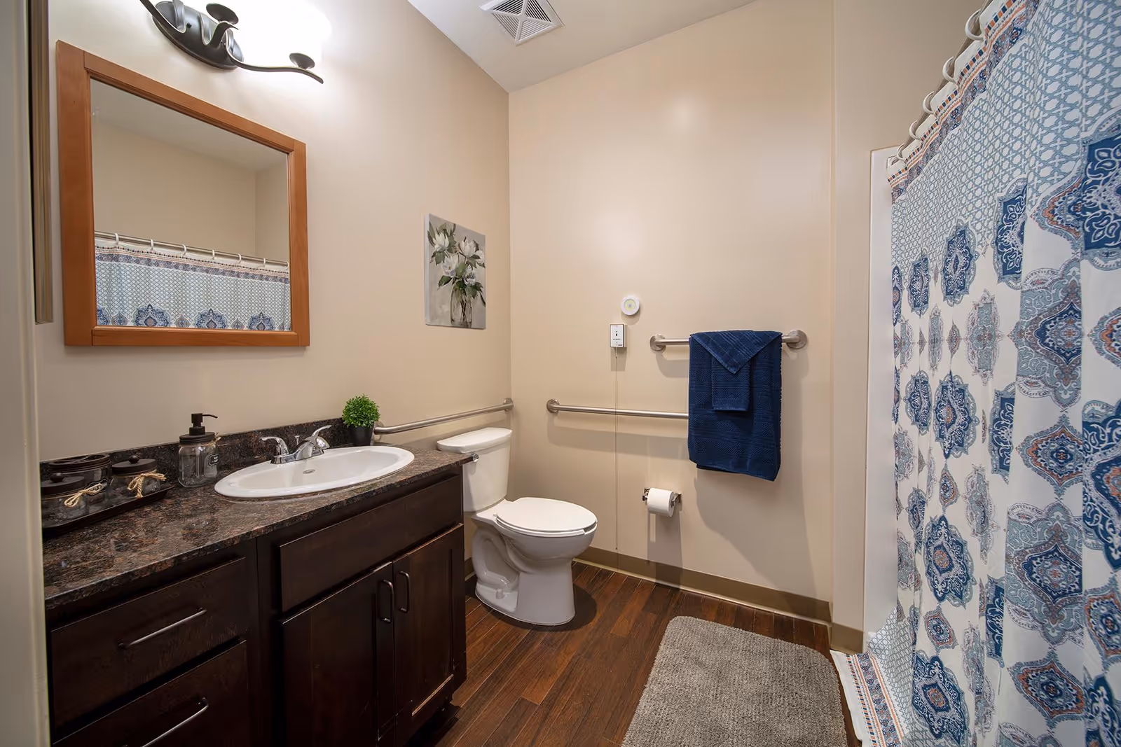 A clean and well-lit bathroom with a dark wood vanity topped with a white sink and a large mirror above it. There is a toilet next to the vanity with a blue towel hanging on a grab bar on the wall. A patterned blue and white shower curtain is visible on the right side, and a gray bath mat is on the wooden floor. A small plant and soap dispenser are on the vanity countertop.
