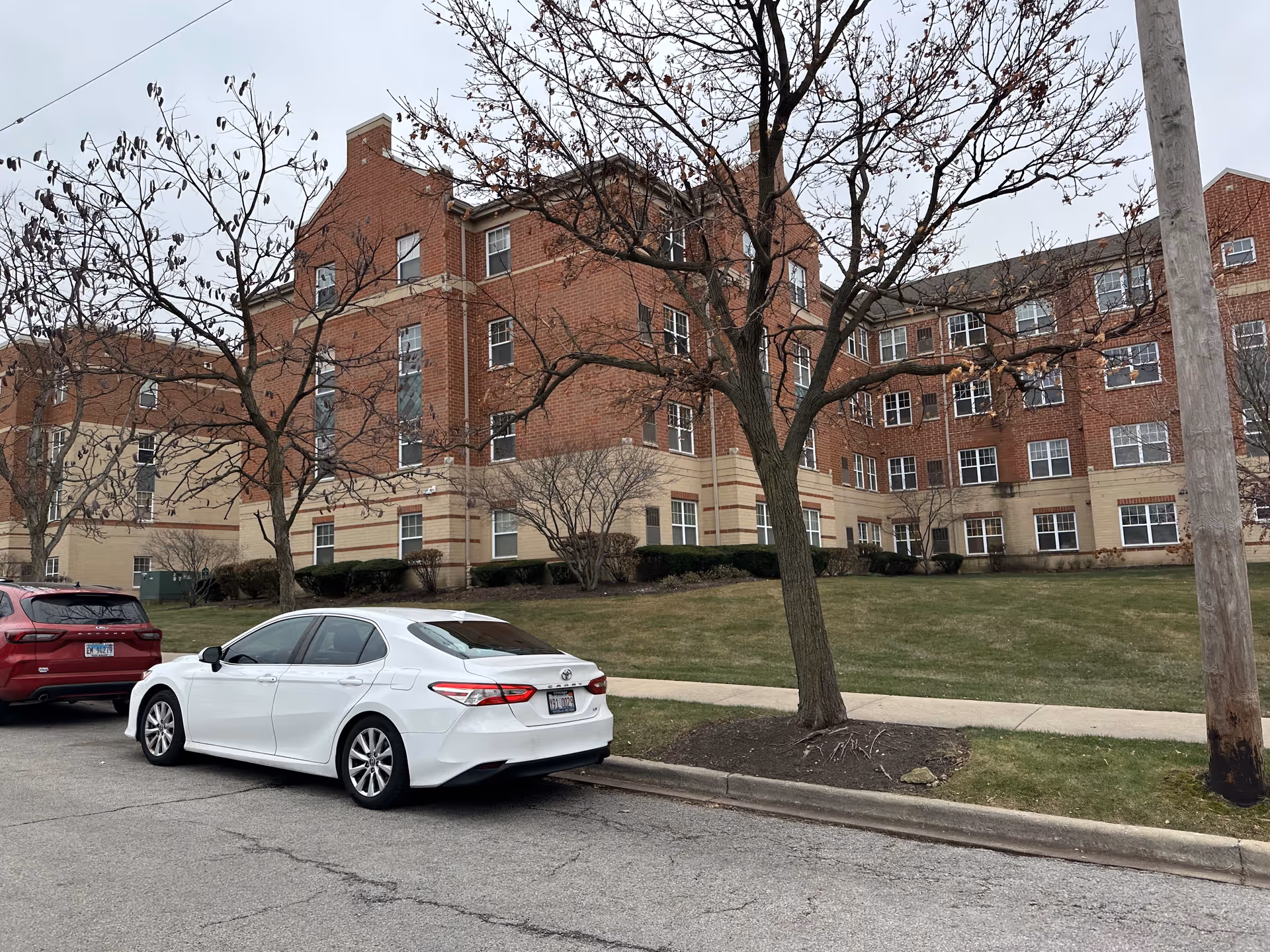 Four-story brick senior living building with a lawn, leafless trees, and parked cars along the street.