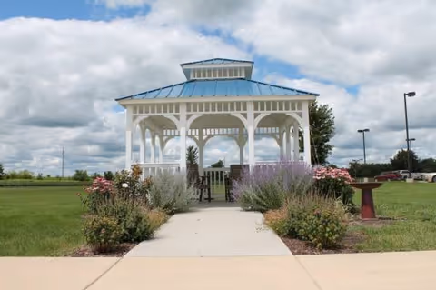 A white gazebo with a blue roof situated at the end of a concrete pathway, surrounded by green grass, flowering plants, and shrubs under a partly cloudy sky.