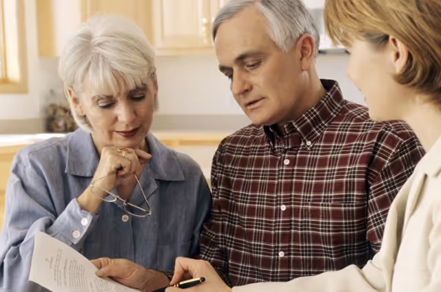 Three adults sitting together indoors, reviewing a document. An older woman with short white hair holds her glasses and looks at the paper, an older man in a plaid shirt points at the document with a pen, and a younger woman in a light-colored blazer listens attentively.