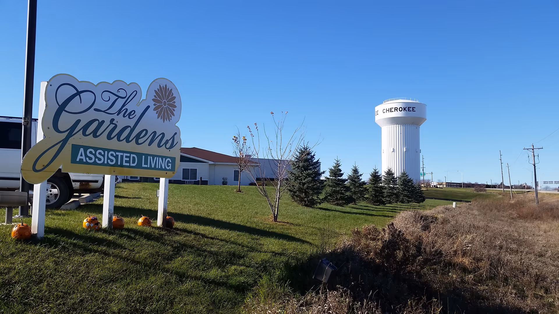 Outdoor view of The Gardens Assisted Living facility sign on a grassy area with a clear blue sky. In the background, there is a white water tower labeled 'CHEROKEE' and some small trees and shrubs.