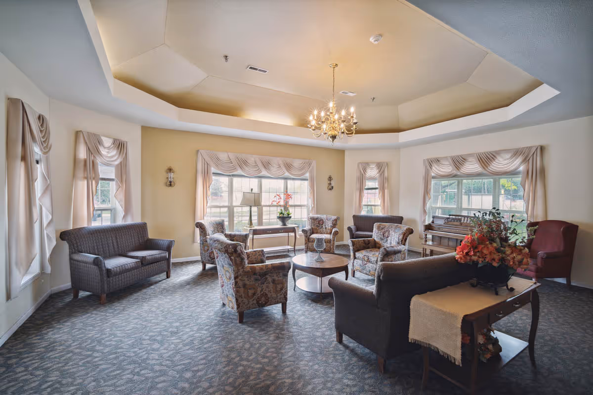 A spacious and well-lit living room with multiple upholstered chairs and sofas arranged around a central wooden coffee table. The room features large windows with draped curtains, a chandelier hanging from a recessed ceiling, a piano near one window, and a side table with a floral arrangement.