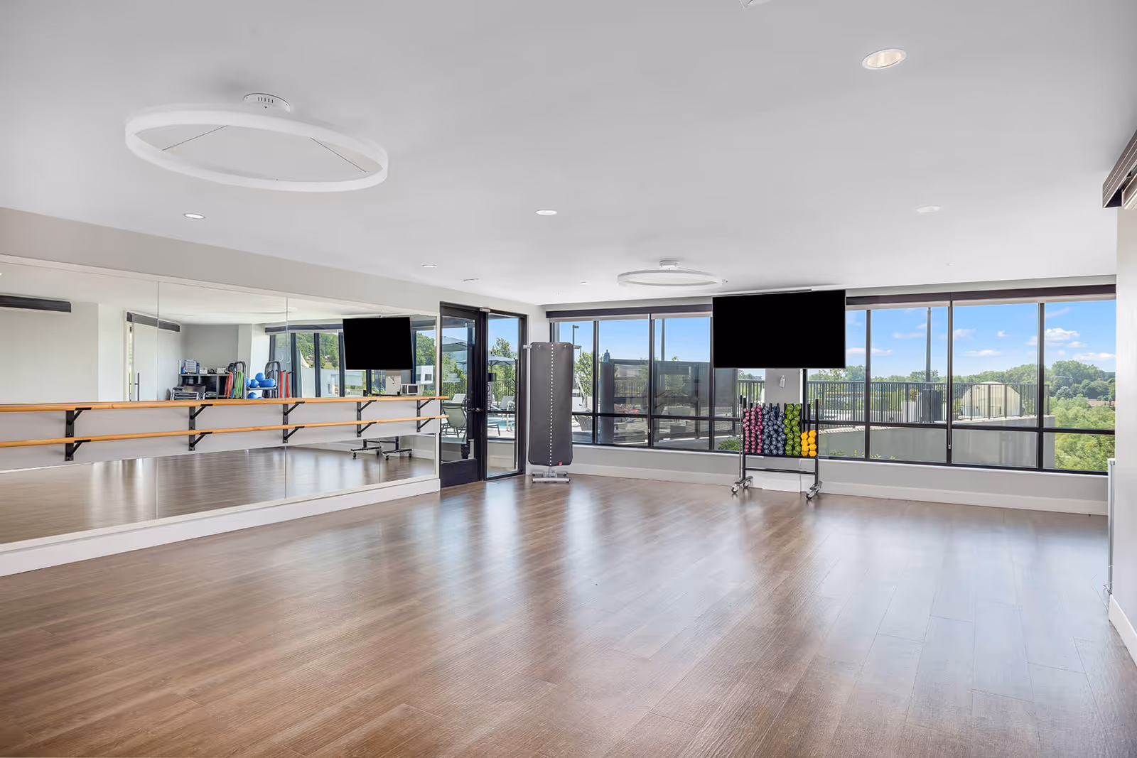 A spacious fitness or exercise room with wooden flooring, large mirrors on one wall with ballet barres, two circular ceiling lights, and large windows providing a view of the outdoors. There is a rack with colorful dumbbells and a black padded exercise mat standing upright near the windows.