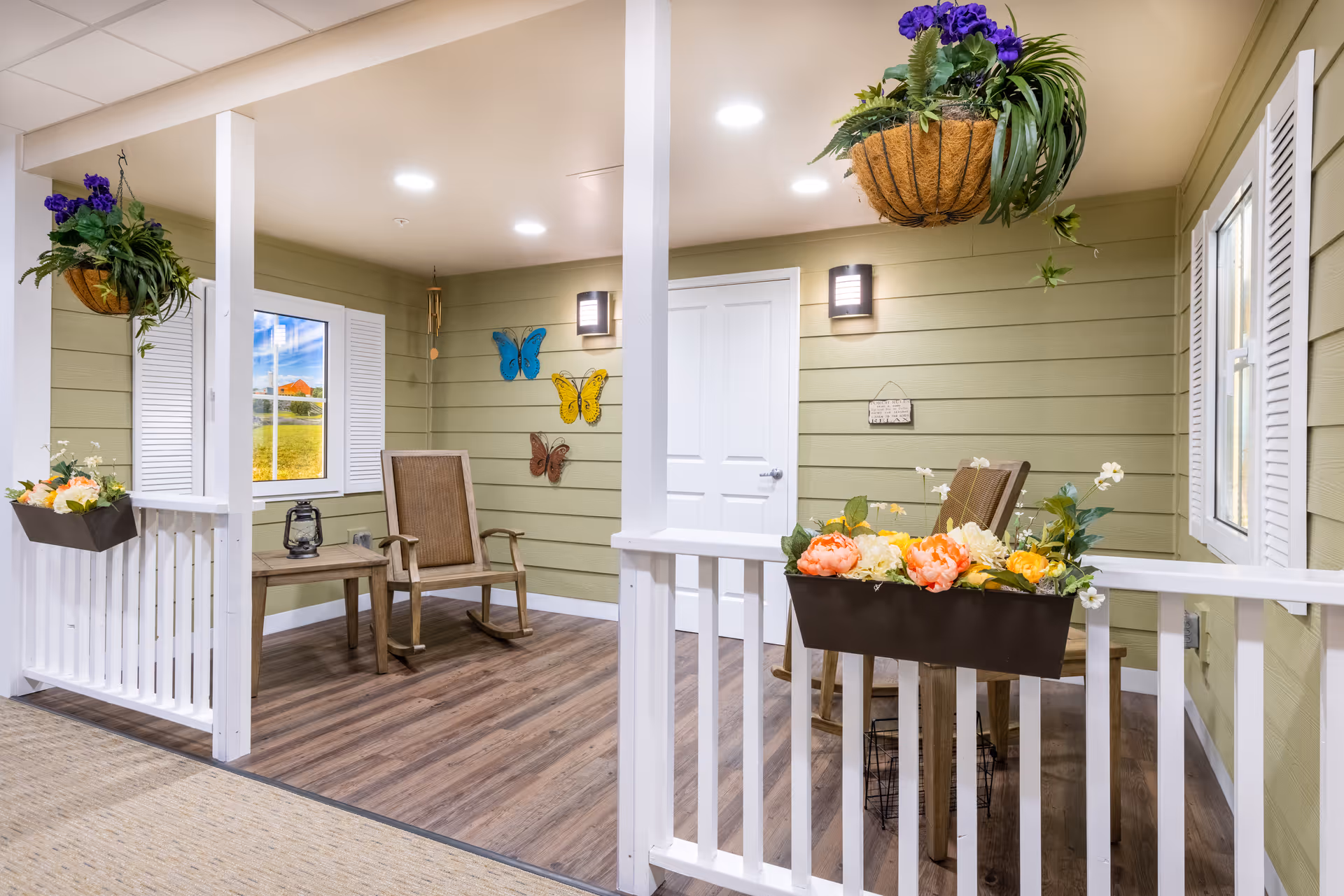 A bright indoor porch-style seating area with wooden rocking chairs, planters of flowers, hanging baskets, and butterfly wall decorations.