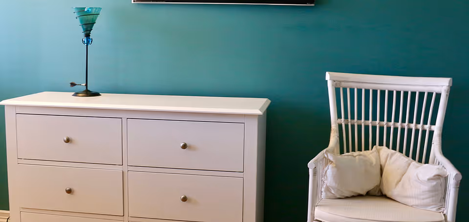 A white dresser with six drawers and a decorative lamp on top is placed against a teal-colored wall. Next to the dresser is a white wicker chair with two white cushions.