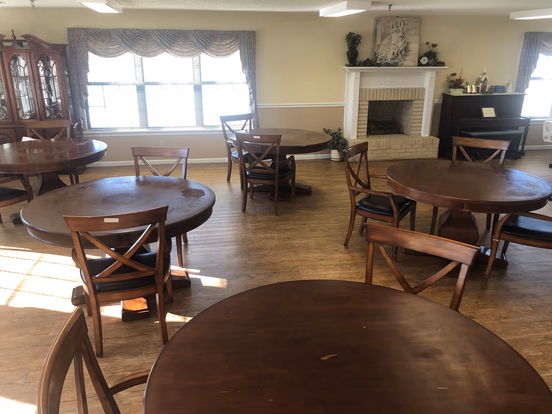 A dining room with several round wooden tables and chairs arranged on a wood floor. There is a large window with patterned curtains letting in natural light, a fireplace with decorative items on the mantel, a piano, and a wooden cabinet with glass doors in the background.