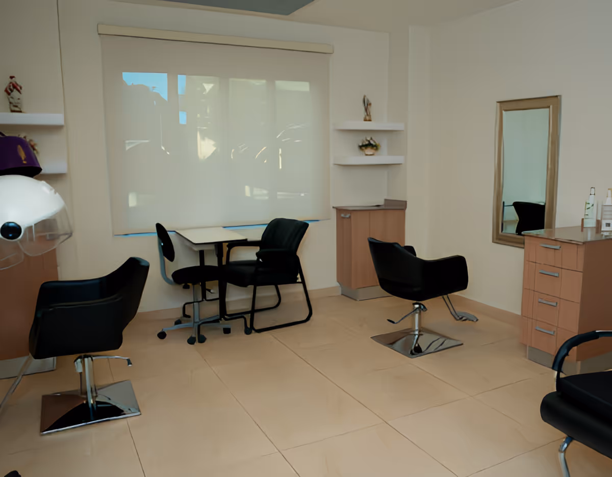 Interior of a salon room with black salon chairs, a hair dryer, a mirror mounted on the wall, a small table with chairs, and shelves with decorative items.