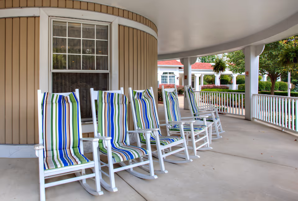 A covered porch area with five white wooden rocking chairs featuring striped cushions in blue, green, white, and brown. The porch has white railings and columns, with greenery and trees visible in the background.