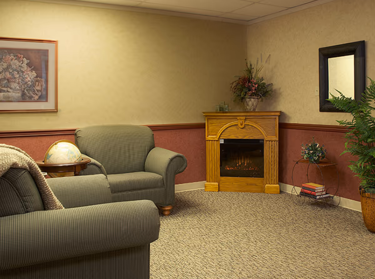 A cozy sitting area with two green upholstered armchairs, a small wooden side table holding a globe, a wooden electric fireplace with a floral arrangement on top, a round metal shelf with books and a small plant, a large potted plant, a framed floral painting on the wall, and a rectangular mirror.
