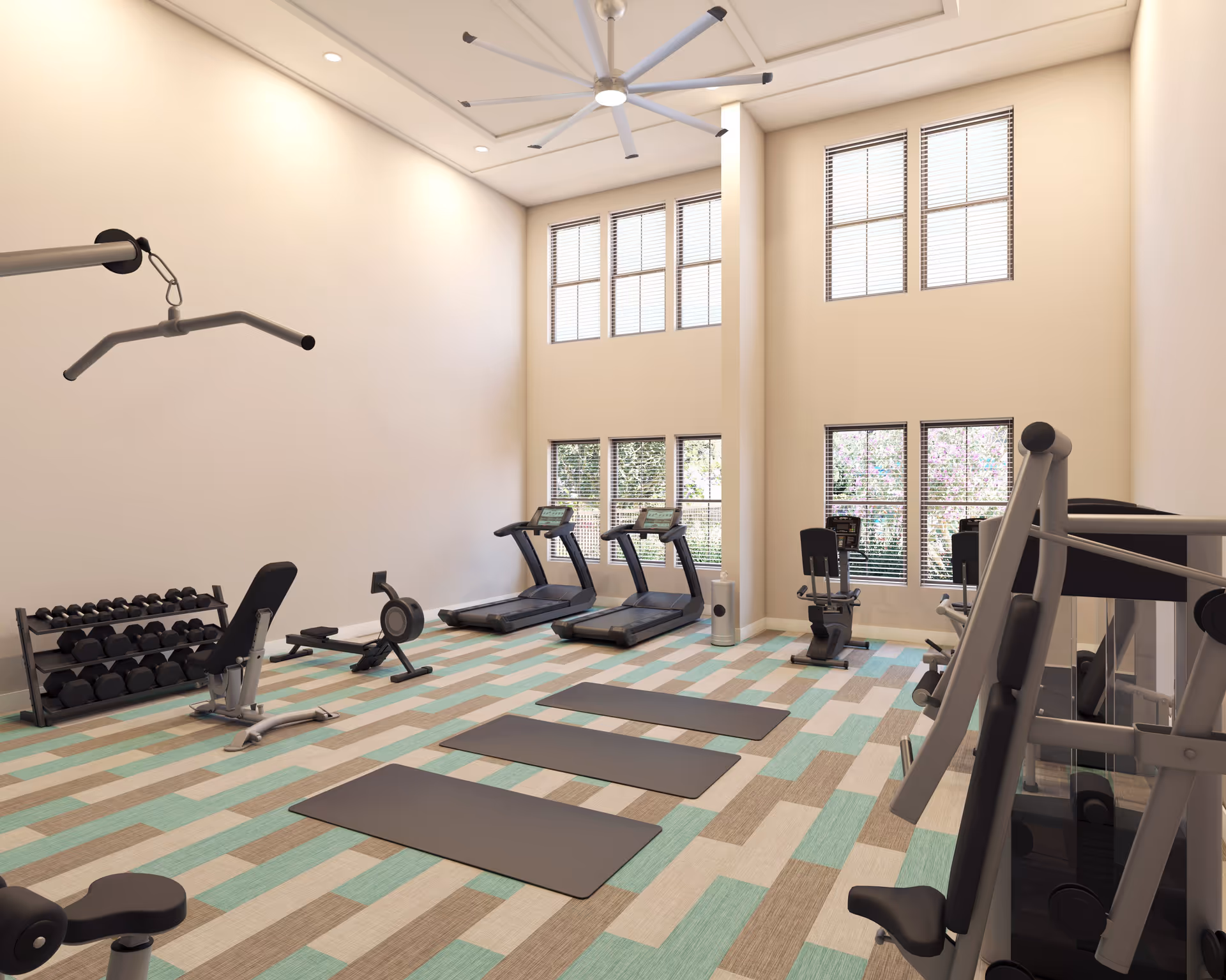 A bright and spacious fitness room with large windows letting in natural light. The room features exercise equipment including treadmills, stationary bikes, a rowing machine, weightlifting bench with dumbbells, and three exercise mats laid out on a patterned floor. A large ceiling fan is mounted above for ventilation.