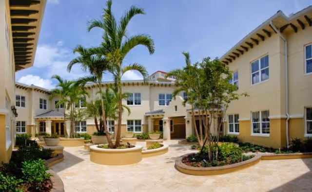 Sunny landscaped courtyard with palm trees and circular planters surrounded by a two-story beige senior living building.