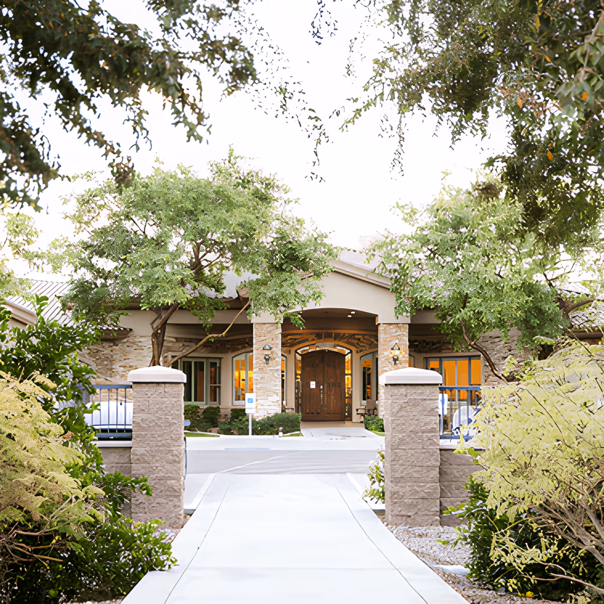 Front entrance of a senior living facility named Emerald Groves Central, featuring a stone pathway leading to a covered porch with large wooden double doors, surrounded by green trees and shrubs.