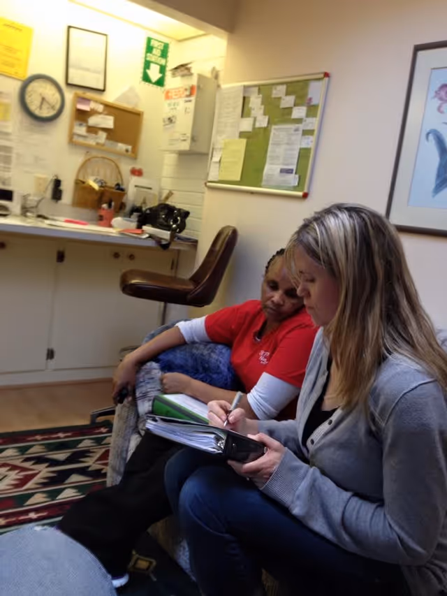 Two women sitting in a small office or common area. One woman in a red shirt is leaning back on a couch, while the other woman with blonde hair is writing in a binder on her lap. The room has a bulletin board with papers, a clock on the wall, and a counter with various items including a basket and a black bag.